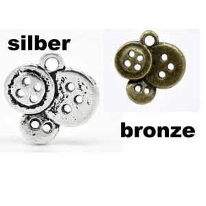 May include: Two button charms, one silver and one bronze, each composed of three buttons of varying sizes. The silver charm has the word "silber" above it. The bronze charm has the word "bronze" below it. Each button has four holes.