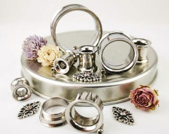 May include: A collection of silver-toned metal ear tunnels and plugs in various sizes, featuring smooth circular designs. Accompanying the jewelry are small, ornate silver-toned filigree charms and dried flowers in shades of purple, cream, and pink.