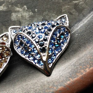 May include: A fox-shaped brooch or pendant featuring a silver-toned metal frame encrusted with numerous small, faceted blue and dark navy crystals. The design highlights the pointed ears and snout of a fox, with a dense, sparkling surface texture.