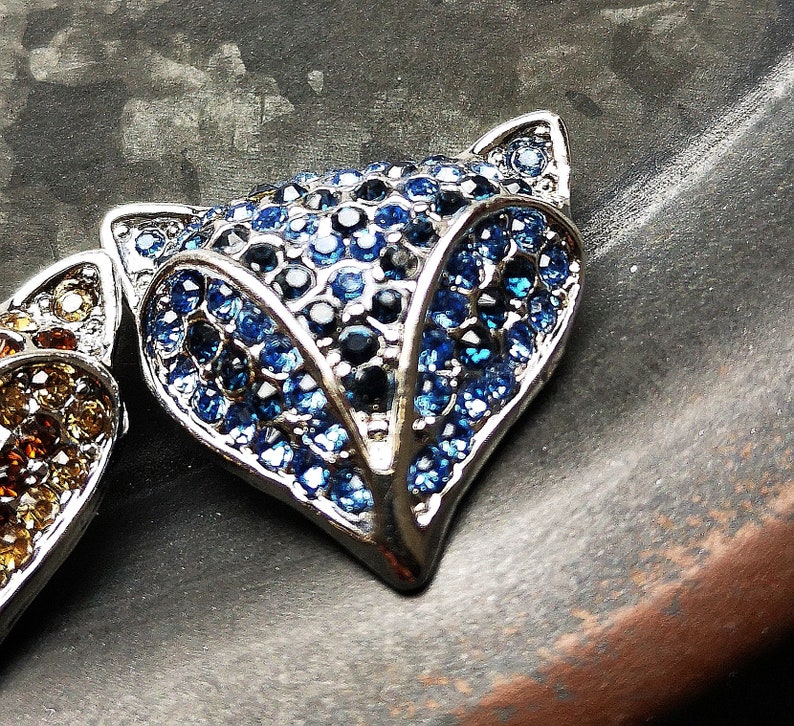 May include: A fox-shaped brooch or pendant featuring a silver-toned metal frame encrusted with numerous small, faceted blue and dark navy crystals. The design highlights the pointed ears and snout of a fox, with a dense, sparkling surface texture.