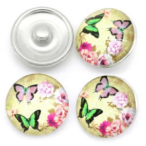 May include: Four circular snap buttons featuring a cream-colored background with a printed pattern of green and pink butterflies alongside clusters of pink and white flowers. Each button has a smooth, rounded surface and a metallic silver-toned edge.
