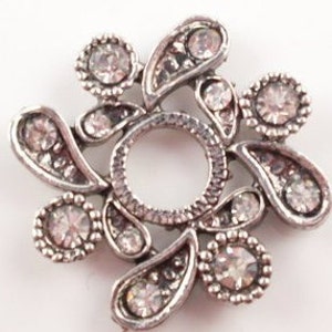 May include: A silver brooch with a floral design. The brooch has a central circular opening with a textured border. Teardrop-shaped and circular elements, each set with clear crystals, form the design. The brooch is set against a white background.