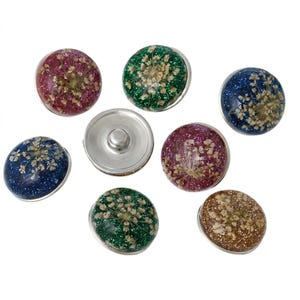 May include: A collection of seven circular snap buttons featuring a domed resin surface. Each button contains embedded dried white flowers and colorful glitter in shades of red, green, blue, purple, and gold. One button shows the silver metal back.