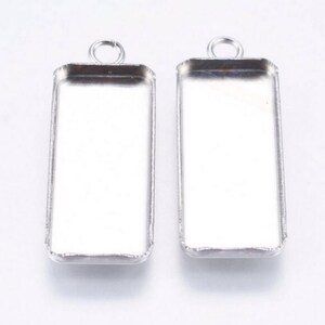 Könnte beinhalten: Zwei silberfarbene Metall-Anhänger-Tabletts mit rechteckiger Form und einer Öse oben. Die Tabletts sind so konzipiert, dass sie einen flach geformten Cabochon oder ein anderes dekoratives Element aufnehmen können.