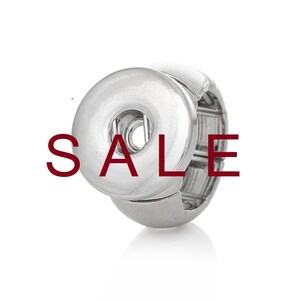 Può includere: Un anello con bottone a pressione argentato con un bottone grande e rotondo. Il bottone ha una superficie liscia e lucida con un piccolo foro al centro.