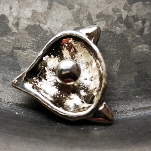 May include: A silver-toned metal brooch shaped like a stylised cat head with pointed ears. The centre of the piece features a smooth, polished spherical bead set against a textured, reflective surface that creates a high-contrast metallic appearance.