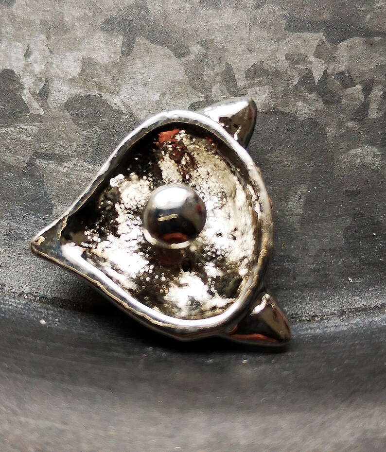 May include: A silver-toned metal brooch shaped like a stylised cat head with pointed ears. The centre of the piece features a smooth, polished spherical bead set against a textured, reflective surface that creates a high-contrast metallic appearance.