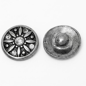Könnte beinhalten: Zwei kreisförmige Metalldruckknöpfe mit antikem Silberfinish. Der vordere Knopf zeigt ein erhabenes Blumenmuster mit eingebetteten klaren Kristallen und kleinen runden Akzenten. Der hintere Knopf hat eine glatte Metalloberfläche mit einem zentralen Zapfen.
