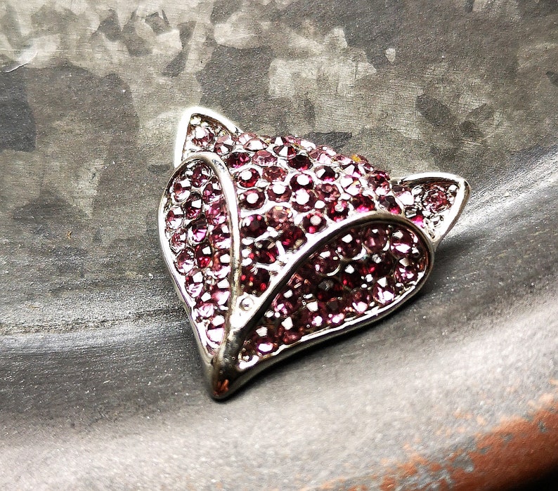 May include: A fox-shaped brooch crafted from silver-toned metal, featuring a dense pav&eacute; setting of small, round, deep pink and magenta crystals that cover the entire surface of the fox head, including the pointed ears and the snout.
