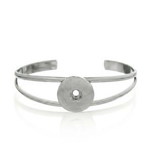 Könnte beinhalten: Ein silberfarbenes Armband aus Metall mit einem doppelten Banddesign, das in der Mitte zusammenläuft. Das Hauptelement ist eine kreisförmige, flache Scheibe mit einem kleinen Loch in der Mitte, das für einen Druckknopfaufsatz gedacht ist.