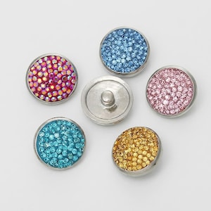 May include: Five circular snap buttons with metallic silver rims. Four buttons feature a surface covered in small, sparkling crystals in shades of pink, light blue, turquoise, and gold. One central button shows the plain silver metal underside.