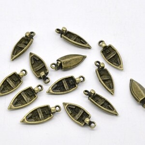 Könnte beinhalten: Eine Sammlung kleiner, antik-messingfarbener Bootsanhänger. Jeder Anhänger hat ein detailliertes Bootsdesign mit Innenplanken und einer Schlaufe zum Befestigen an Schmuck oder anderen Bastelarbeiten. Die Anhänger sind auf einer weißen Oberfläche verstreut.
