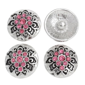 May include: Four silver-toned snap buttons with a floral design. Each button features a central cluster of pink rhinestones surrounded by an ornate, swirling pattern. The reverse side of one button is visible.