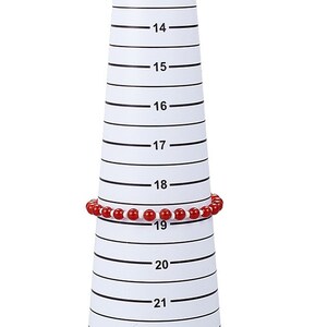 Könnte beinhalten: Ein weißer Kunststoff-Ringgrößenmesser mit schwarzen Markierungen in Zentimetern von 12 bis 22. Ein rotes Perlenarmband ist auf dem Größenmesser an der 19 cm-Marke platziert.