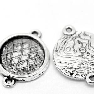 May include: Two silver metal jewellery components. One has a round frame with a grid pattern, the other a textured, organic design. Both have loops for attachment, suitable for crafting.