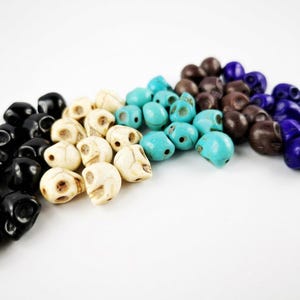 May include: A collection of small skull-shaped beads arranged in a curved line. The beads are colored in black, off-white, bright turquoise, dark brown, and deep blue. Each bead features carved eye sockets and a nasal cavity, creating a detailed skull form.