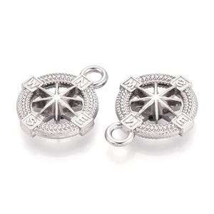 Pode incluir: Dois pingentes prateados em forma de bússola. Cada pingente apresenta um design de rosa dos ventos com letras direcionais (N, S, E, O) e uma borda texturizada. Os pingentes têm um pequeno laço para fixação.