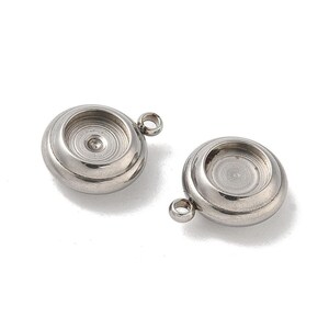 May include: Two stainless steel bezel cups with a circular design. Each cup has a recessed center and a small loop for attaching to a chain or other jewelry component. The cups are on a white background.