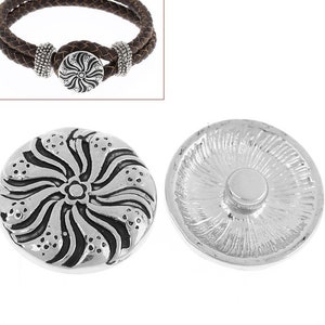 May include: A silver-toned circular snap button featuring a raised, swirling floral pattern with dark recessed accents. The reverse side shows a smooth, flat circular attachment post. A brown braided leather bracelet is shown for context.