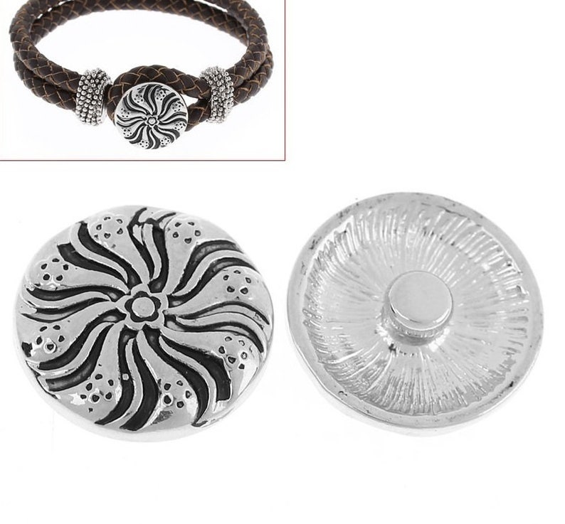 May include: A silver-toned circular snap button featuring a raised, swirling floral pattern with dark recessed accents. The reverse side shows a smooth, flat circular attachment post. A brown braided leather bracelet is shown for context.