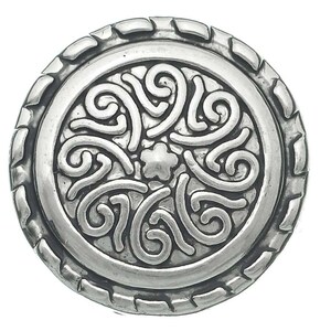 May include: A circular silver-toned button featuring a central star-shaped motif surrounded by raised, swirling scrollwork patterns. The outer edge of the button is defined by a textured, notched border that frames the intricate, embossed design.