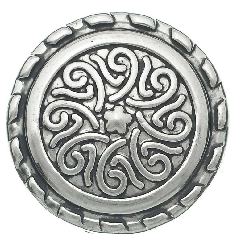 May include: A circular silver-toned button featuring a central star-shaped motif surrounded by raised, swirling scrollwork patterns. The outer edge of the button is defined by a textured, notched border that frames the intricate, embossed design.