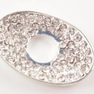 May include: A silver oval charm with a circle cut out in the centre. The charm is covered in small, clear rhinestones.