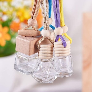 Könnte beinhalten: Klare Glas-Lufterfrischerflaschen für Autos mit Holzaufsätzen und bunten Kordeln. Die Flaschen haben verschiedene geometrische Formen. Die Kordeln sind beige, blau, lila und gelb. Der Hintergrund ist mit Blumen verschwommen.