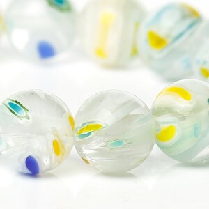 May include: A strand of clear, round glass beads with a white base and colorful accents. The beads feature yellow, blue, and green swirls and dots. The beads are approximately 0.75 inches in diameter.
