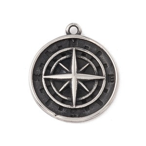 Pode incluir: Um pingente de prata em forma de bússola com fundo preto e uma rosa dos ventos prateada. A rosa dos ventos tem quatro pontos e é cercada por um círculo com as letras N, E, S e W.