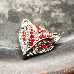 May include: A fox head brooch crafted from silver-toned metal, featuring a dense arrangement of sparkling clear and vibrant red rhinestones. The design highlights the pointed ears and facial features of a fox, with the stones set in a metallic frame.