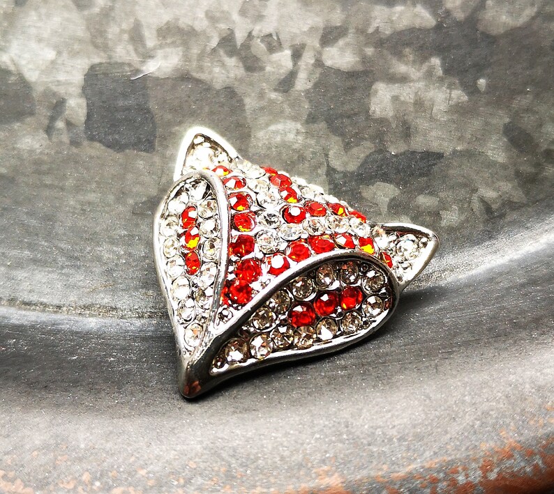 May include: A fox head brooch crafted from silver-toned metal, featuring a dense arrangement of sparkling clear and vibrant red rhinestones. The design highlights the pointed ears and facial features of a fox, with the stones set in a metallic frame.