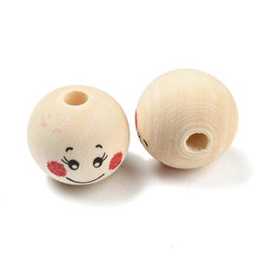 May include: Two wooden beads with painted faces. Each bead is round, with a hole drilled through the centre. One bead has a smiling face with black eyes and red cheeks. The other bead has a partial face.