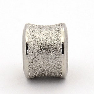 Könnte beinhalten: Eine silberfarbene Metallperle mit strukturierter Oberfläche. Die Perle ist zylindrisch geformt und hat ein leicht gebogenes Design.