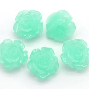 May include: Five light green, translucent rose-shaped beads. Each bead has detailed petals and a rounded shape. The beads are arranged on a white background, showcasing their delicate floral design. Ideal for jewelry making or craft projects.