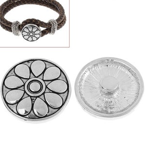 Pode incluir: Um bot&atilde;o de press&atilde;o de metal prateado com um design de p&eacute;talas florais em relevo na frente e um fecho circular na parte de tr&aacute;s, exibido ao lado de uma pulseira de couro tran&ccedil;ado castanho com contas prateadas e fecho de press&atilde;o a condizer.