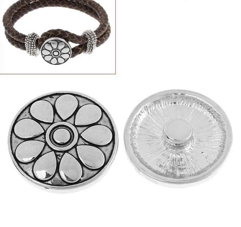 Pode incluir: Um bot&atilde;o de press&atilde;o de metal prateado com um design de p&eacute;talas florais em relevo na frente e um fecho circular na parte de tr&aacute;s, exibido ao lado de uma pulseira de couro tran&ccedil;ado castanho com contas prateadas e fecho de press&atilde;o a condizer.