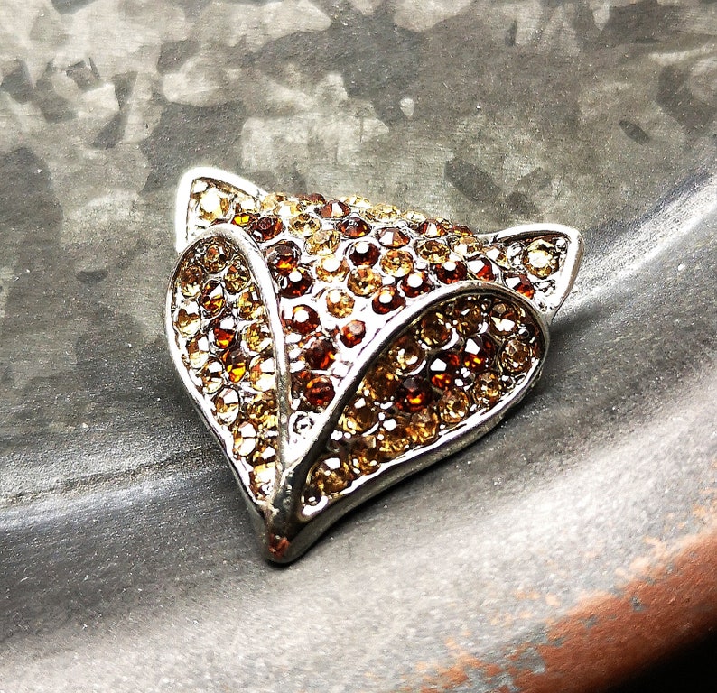 May include: A fox head brooch crafted from silver-toned metal, featuring a dense arrangement of faceted crystals in shades of amber, copper, and deep brown. The design captures the pointed ears and snout of a fox, with the stones creating a textured surface.