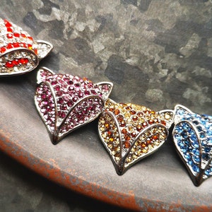 May include: A necklace featuring four fox head pendants, each encrusted with small sparkling crystals. The pendants are arranged in a row, displaying distinct colour variations of red and white, deep pink, golden yellow, and bright blue.
