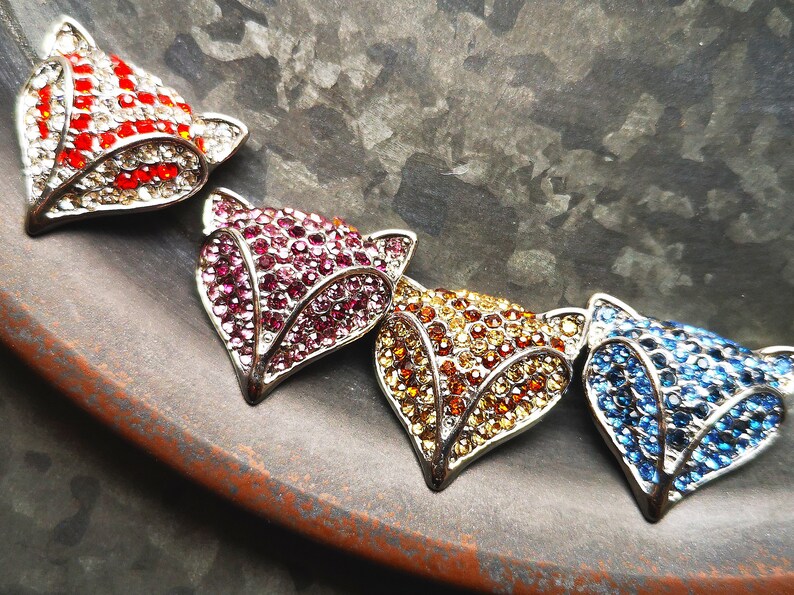 May include: A necklace featuring four fox head pendants, each encrusted with small sparkling crystals. The pendants are arranged in a row, displaying distinct colour variations of red and white, deep pink, golden yellow, and bright blue.