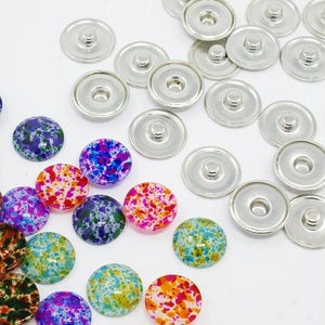 Könnte beinhalten: Eine Sammlung von silberfarbenen Metall-Druckknöpfen und bunten, gesprenkelten Harz-Cabochons. Die Cabochons weisen eine Vielzahl von Farben auf, darunter Blau, Lila, Grün, Orange und Rosa, mit einem gesprenkelten Muster. Die Verschlüsse sind rund.