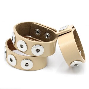 Könnte beinhalten: Drei beige Lederarmbänder mit silberfarbenen Druckknöpfen. Die Armbänder sind auf weißem Hintergrund gestapelt. Das Leder ist glatt und die Druckknöpfe sind gleichmäßig verteilt. Die Armbänder sind etwa 2 cm breit.
