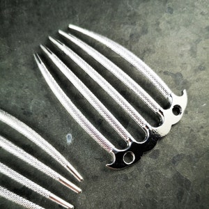 Könnte beinhalten: Silberner Haarkamm mit dekorativem Design und fünf Zähnen. Der Kamm ist aus Metall und hat eine glänzende Oberfläche.