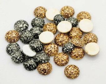 May include: A collection of small, round, dome-shaped cabochons featuring a speckled, marbled pattern in shades of black, white, and tan. Some pieces show the smooth, flat, solid white underside, while others display the textured, patterned top surface.