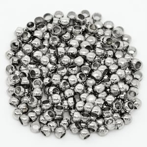 May include: A large collection of small, round, metallic silver beads with a polished, reflective finish. Each bead features a central hole, creating a uniform appearance across the pile. The beads are arranged in a dense, circular cluster.