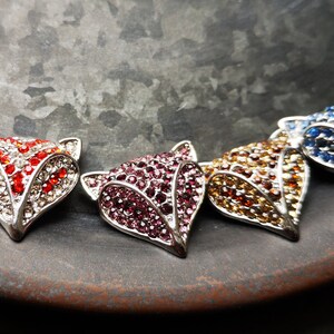 May include: Four fox-shaped metal charms, each encrusted with small sparkling rhinestones in distinct colours: red and white, deep pink, amber, and blue. The fox heads feature pointed ears and a triangular snout, with a metallic silver-toned frame.
