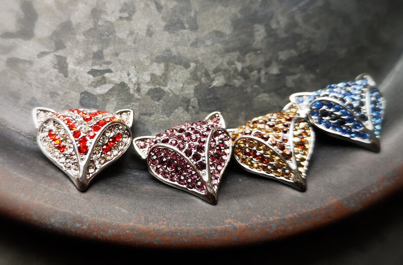 May include: Four fox-shaped metal charms, each encrusted with small sparkling rhinestones in distinct colours: red and white, deep pink, amber, and blue. The fox heads feature pointed ears and a triangular snout, with a metallic silver-toned frame.