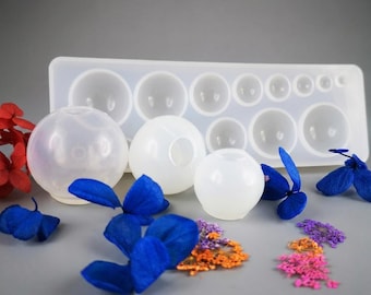 Puede incluir: Un molde de silicona translúcido con múltiples cavidades hemisféricas de varios tamaños, acompañado de tres objetos esféricos de resina con aberturas circulares, rodeados de flores secas azules, rojas, naranjas y moradas esparcidas.
