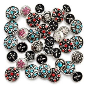 Könnte beinhalten: Eine Sammlung dekorativer Knöpfe in verschiedenen Designs. Einige Knöpfe haben eine silberfarbene Metallbasis mit türkisfarbenen, roten oder rosa Edelsteinen. Andere Knöpfe haben eine schwarze Emaille-Oberfläche mit einem Fleur-de-Lis-Design. Die Knöpfe sind rund und unterschiedlich groß.