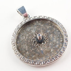 Può includere: Ciondolo argentato con design circolare. Presenta un bordo decorativo con un motivo a chiave greca. Il centro ha una superficie strutturata con una vite e un piccolo anello di gomma nero. Il passante è di forma rettangolare semplice.